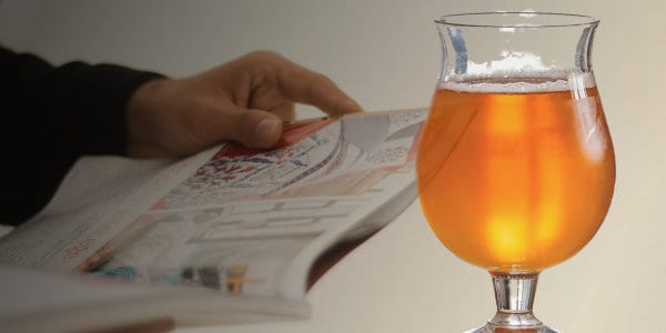 Glass of beer with person holding magazine in background.