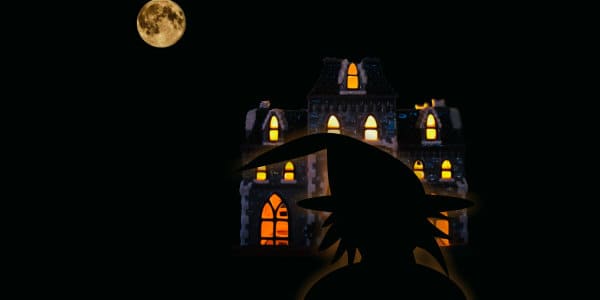 Witch silhouette in front of spooky house with moon.