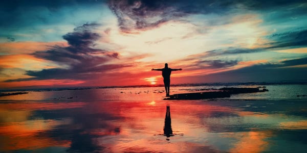 Person standing with arms stretched on a beach at sunrise with red coloured sky reflected in the water.